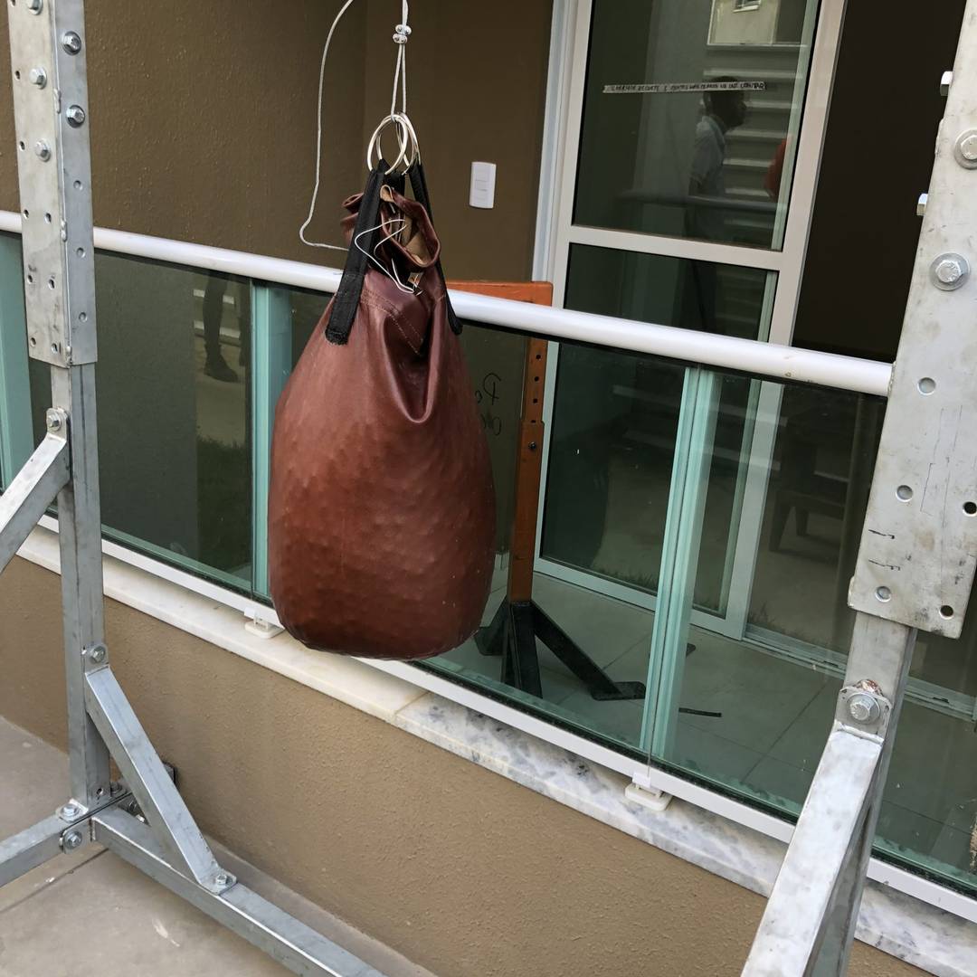Saco de couro do ensaio de impacto de corpo mole suspenso no pórtico metálico, posicionado diante do guarda-corpo de vidro da varanda.
