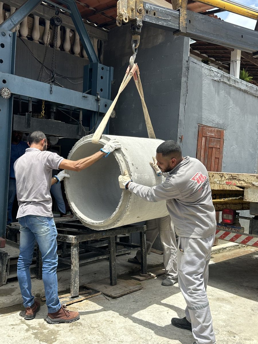 Dois técnicos içando um tubo de concreto por cinta de nylon para posicioná-lo no pórtico azul da prensa, antes do ensaio de compressão.