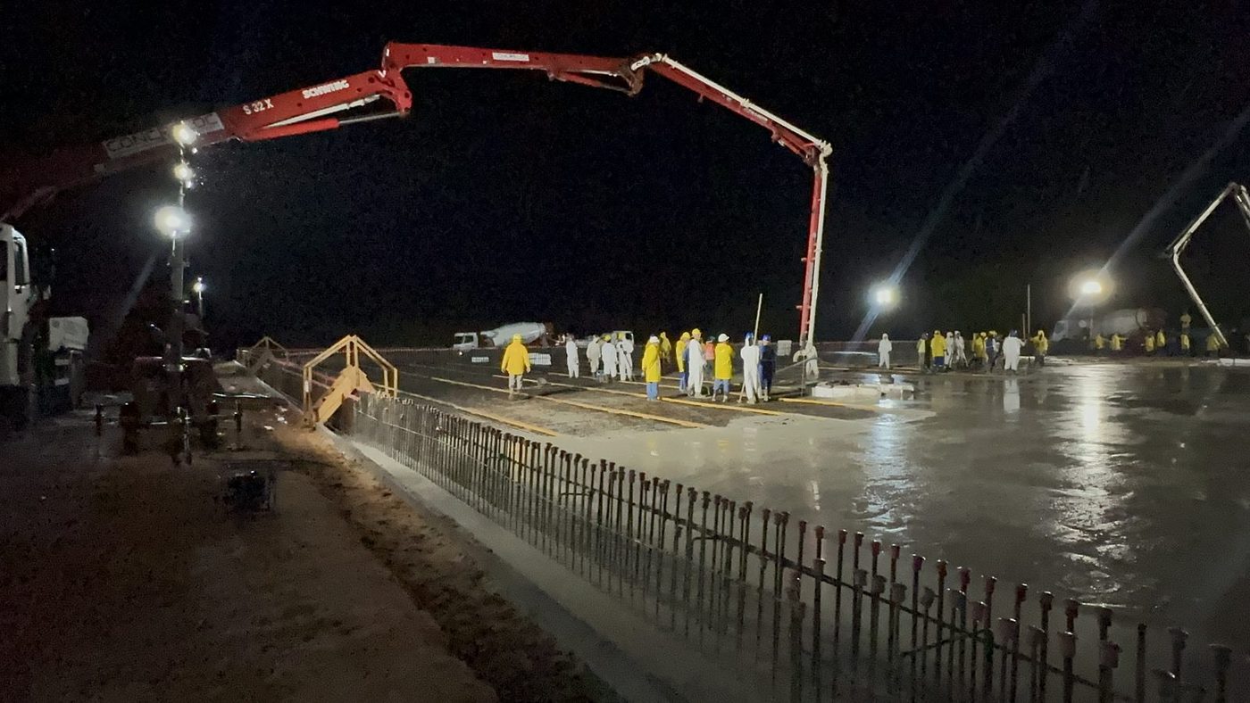 Concretagem noturna de laje com bomba lança em operação e equipe de macacão distribuída pela frente de serviço, sob iluminação de canteiro.