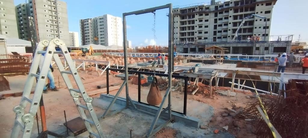 Pórtico metálico do ensaio de impacto de corpo mole montado em canteiro, com o saco suspenso por cabo e escada de apoio ao lado; edifícios em construção ao fundo.