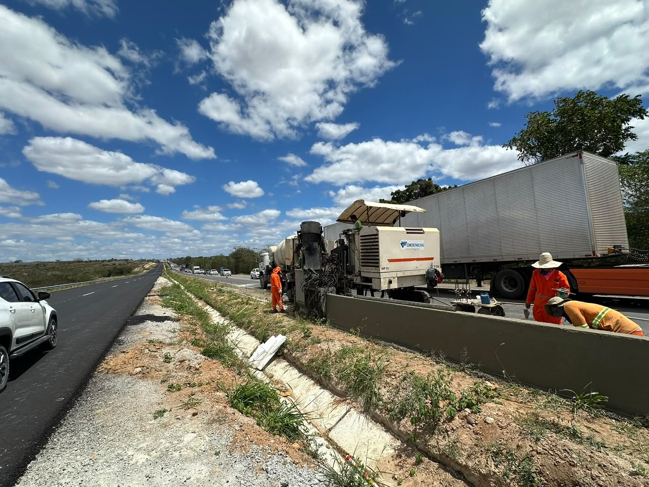 Máquina de fôrma deslizante executa barreira New Jersey na lateral de rodovia, com caminhão-betoneira e equipe uniformizada.