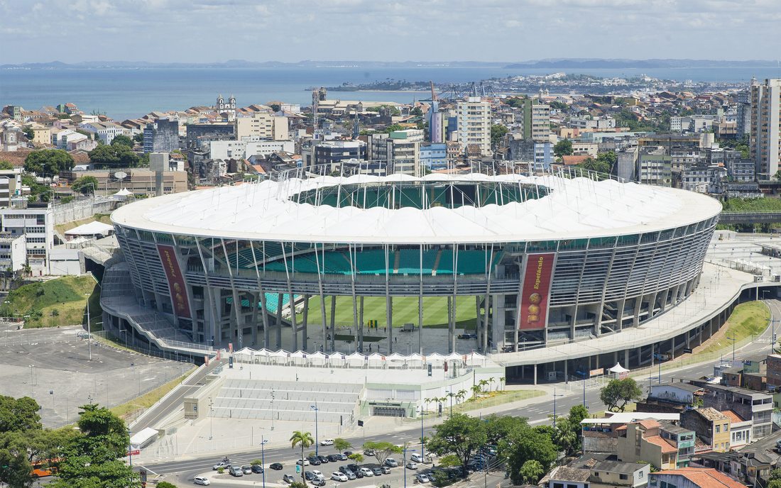 Vista aérea da Itaipava Arena Fonte Nova, em Salvador-BA, com a cobertura circular e a cidade e o mar ao fundo — inspeção dos pilares de concreto e ensaios especiais.