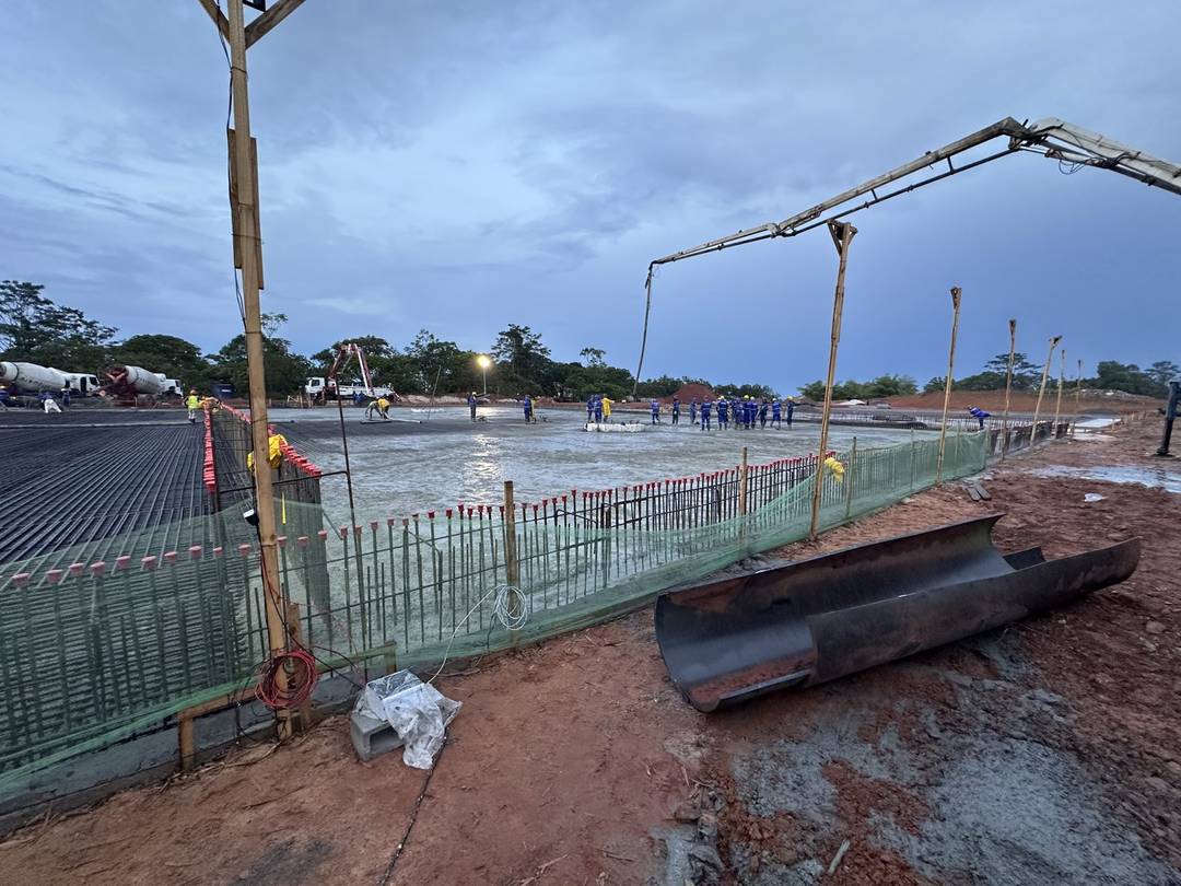 Vista ampla de canteiro ao entardecer: laje em concretagem cercada por tela de proteção, bomba lança em operação e equipe distribuída pela frente de serviço.