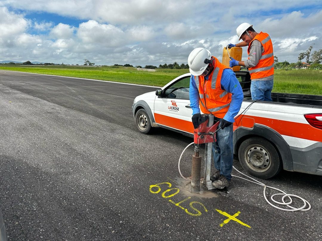 Dois técnicos da LACROSE de colete refletivo extraindo testemunho de pavimento asfáltico com extratora rotativa, ao lado da caminhonete da empresa e da marcação ST09 em tinta amarela.