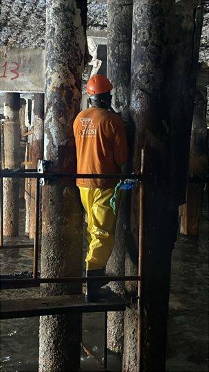 Operário em recuperação estrutural de estacas de concreto, trabalhando entre pilares sobre plataforma de madeira.