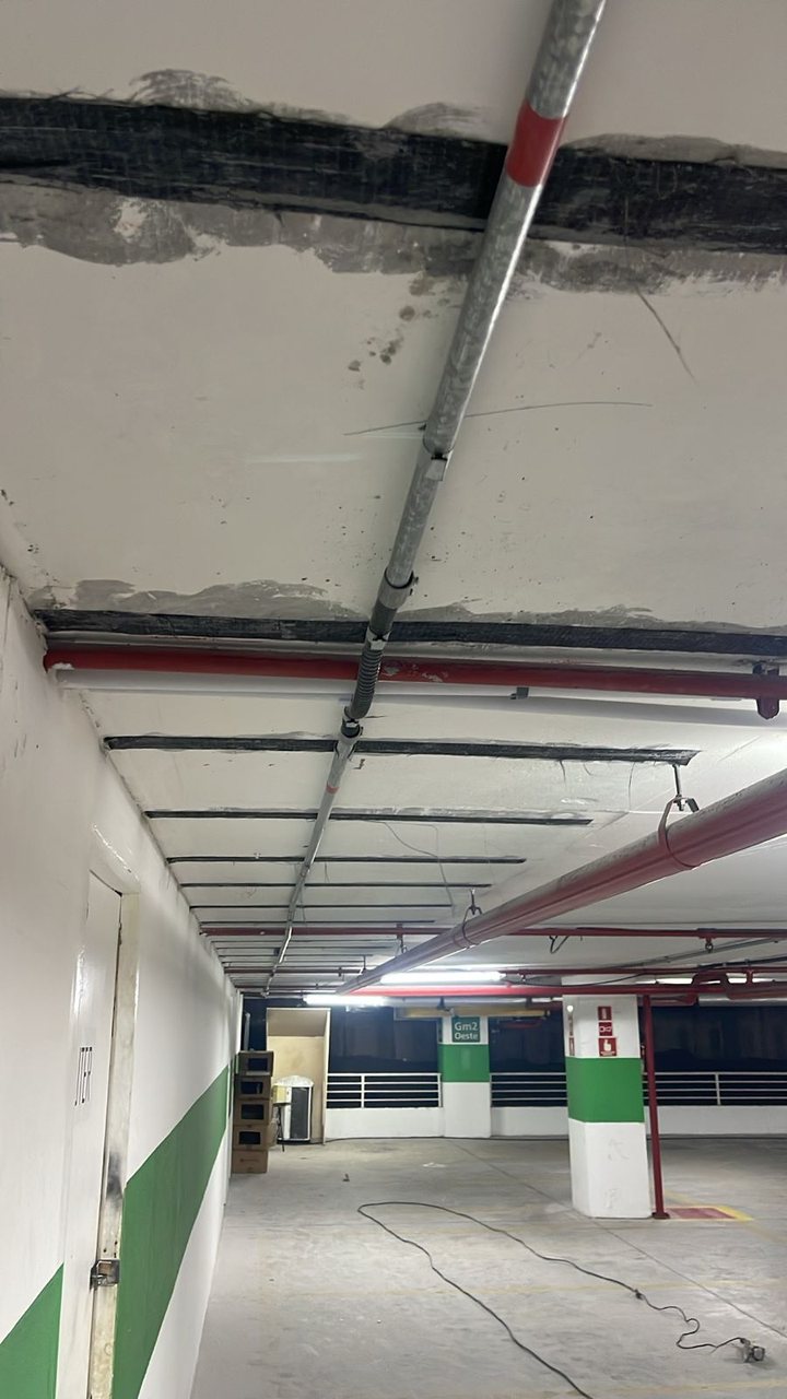 Teto de garagem com laminados de fibra de carbono colados à face inferior da laje, formando faixas escuras paralelas entre as tubulações.
