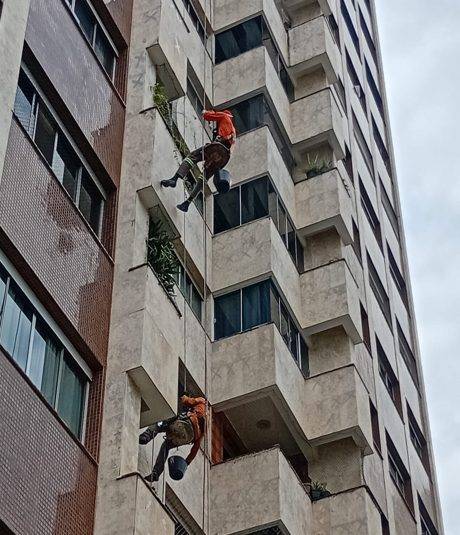 Fachada de edifício residencial em obra de retrofit, com dois profissionais em acesso por corda recuperando o revestimento.